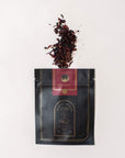 A black and gold package labeled "Magic Hour" with a red section labeled "Moon Lover: Herbal Immunitea Ritual Gift" is open at the top, spilling its organic loose leaf tea onto a white background. The dark tea leaves display a rich mix of red and brown hues.