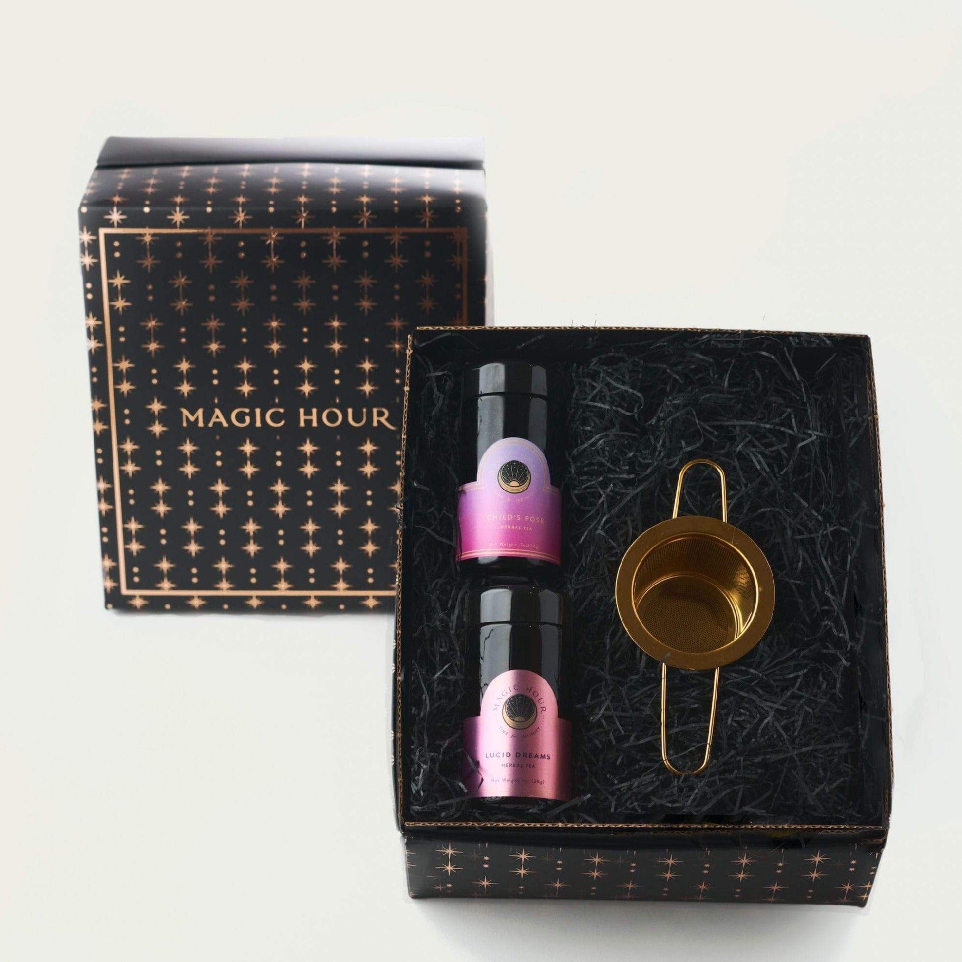 A luxury gift set featuring two small bottles of essential oils and a gold-toned tea strainer. The set is packaged in a black box adorned with a starry pattern and the words "Magic Hour" on the lid. The bottles have pink and purple labels. Perfect for the organic tea enthusiast in your life, this Drift into Sleep Ritual Gift will delight their senses.