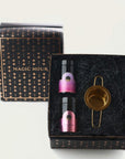A luxury gift set featuring two small bottles of essential oils and a gold-toned tea strainer. The set is packaged in a black box adorned with a starry pattern and the words "Magic Hour" on the lid. The bottles have pink and purple labels. Perfect for the organic tea enthusiast in your life, this Drift into Sleep Ritual Gift will delight their senses.