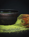 A traditional bamboo whisk sits beside a dark ceramic bowl filled with matcha tea. The Artisanal Magic Hour Matcha Whisk by Magic Hour is nestled within, while matcha powder is sprinkled around them on a dark surface, highlighting the vibrant green hue of this organic tea.