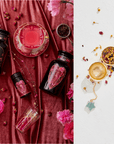 An overhead view of the Cosmic Gemstone Apothecary Gift Set showcases an opulent display with black heart-adorned bottles and a ruby red drink in an ornate glass on rich velvet fabric. To the right, loose tea and a gold Emerald Star Tea Strainer glitter on a pristine white surface alongside vibrant dried flowers, all surrounded by delicate pink blossoms.