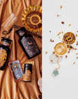 An elegant arrangement featuring black and gold tea containers, a tarot card, and a string of black beads on the left side. The right side highlights the Emerald Star Tea Strainer from the Cosmic Gemstone Apothecary Gift Set with dried flowers and loose leaf tea scattered on a white surface.
