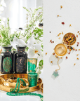 A split image with the left side showcasing various glass bottles, jewelry, and a golden cup arranged on a golden tray adorned with white flowers and greenery. The right side features an arrangement of dried tea leaves, an Astrology Tea infuser, and scattered flowers from the Cosmic Gemstone Apothecary Gift Set.
