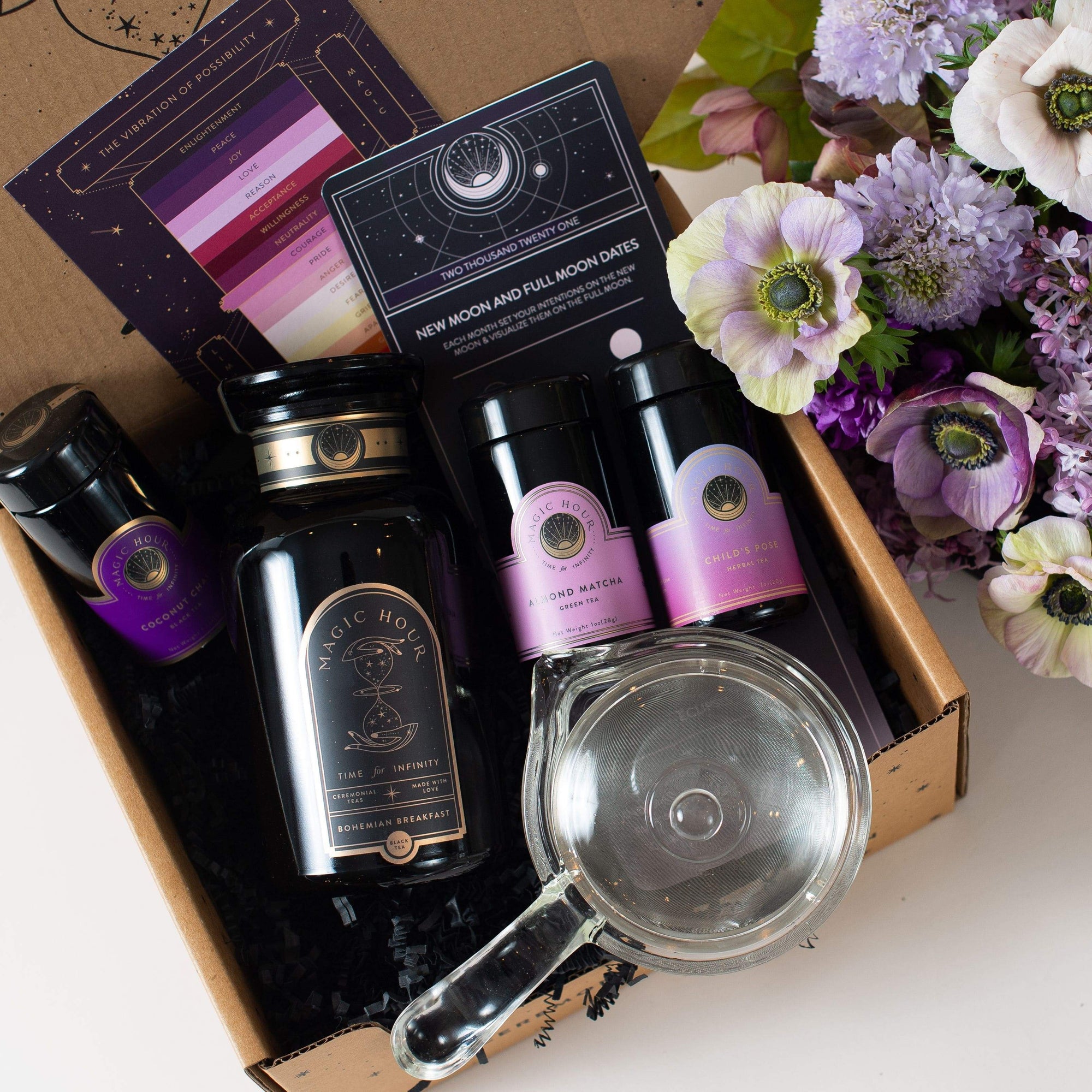 A flat-lay image of Zhena's Essential Favorites Tea Collection gift box, featuring a bottle of "Magic Hour" tea, two small jars of Almond Matcha tea, a tea infuser, a booklet, and some flowers on the side. The items are arranged neatly against a background of crinkled black paper. A bouquet of pink and purple flowers is on the right.