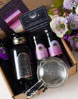 A flat-lay image of Zhena's Essential Favorites Tea Collection gift box, featuring a bottle of "Magic Hour" tea, two small jars of Almond Matcha tea, a tea infuser, a booklet, and some flowers on the side. The items are arranged neatly against a background of crinkled black paper. A bouquet of pink and purple flowers is on the right.