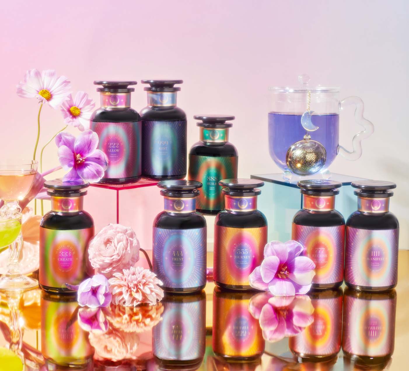 A colorful display of iridescent bottles surrounded by pink and purple flowers. A glass of blue liquid with a tea strainer sits in the background. The scene is set against a pastel backdrop.