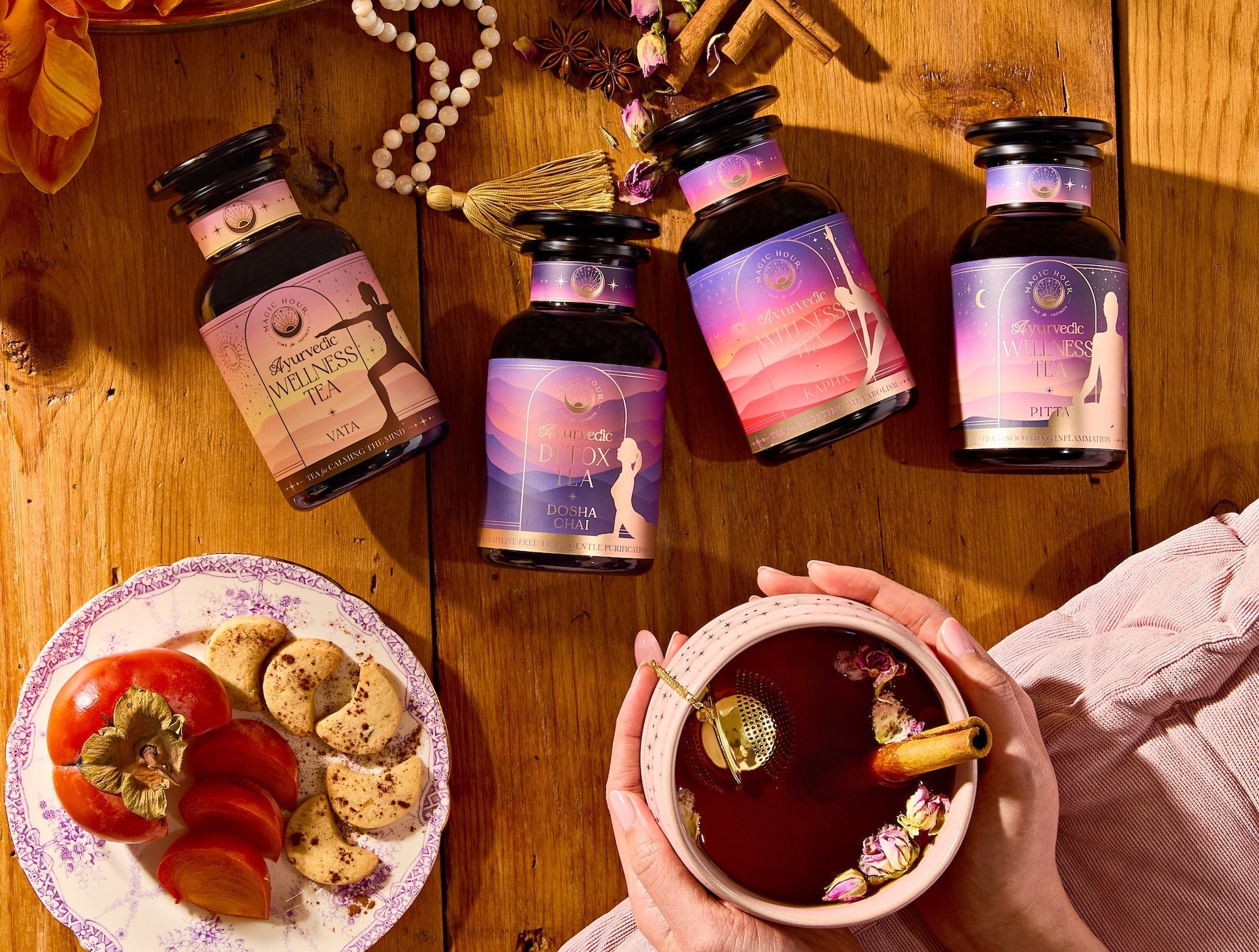 Four jars of wellness tea on a wooden table, a hand holding a cup of tea with a gold stick, and a plate with cookies and a persimmon, surrounded by autumn decor and beads.