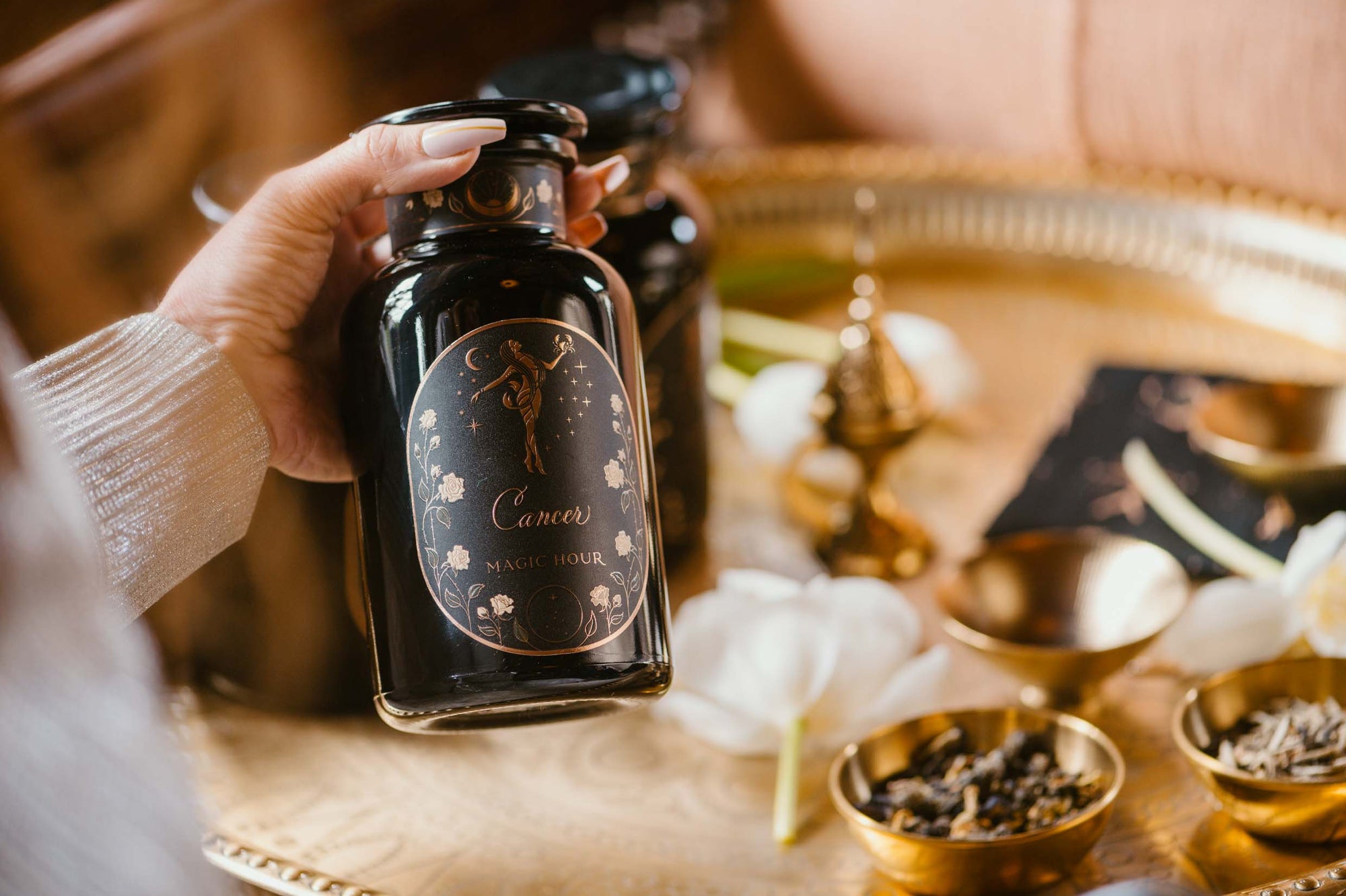 A hand holds a black glass jar labeled "Cancer Magic Hour" over a gold tray with gold bowls, loose tea leaves, white flower petals, and an incense burner.