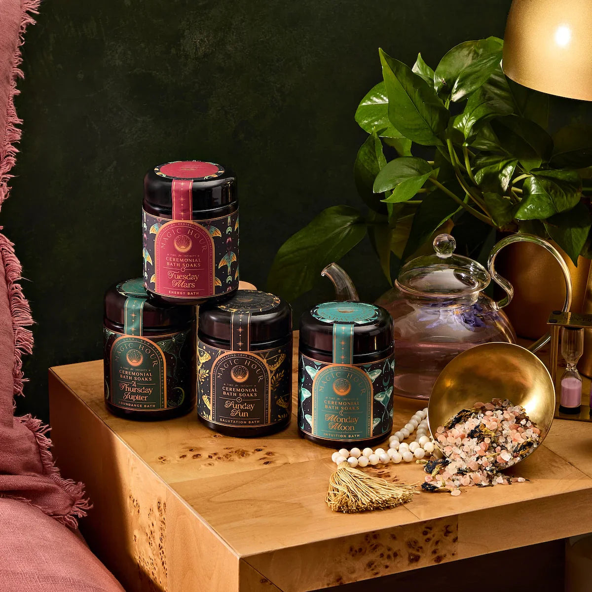 Four decorative jars of wellness powders are displayed on a wooden table next to a green plant, a glass teapot, a gold scoop with bath salts, and a pink couch with a tasseled cushion.