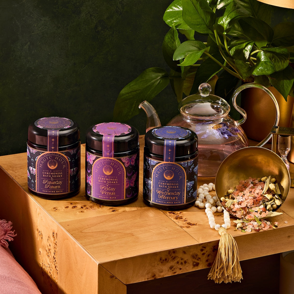 Three jars of herbal tea are placed on a wooden table. Each jar has a different astrological sign label. Nearby, a teapot and a gold scoop with loose tea are displayed. Green leaves are in the background.