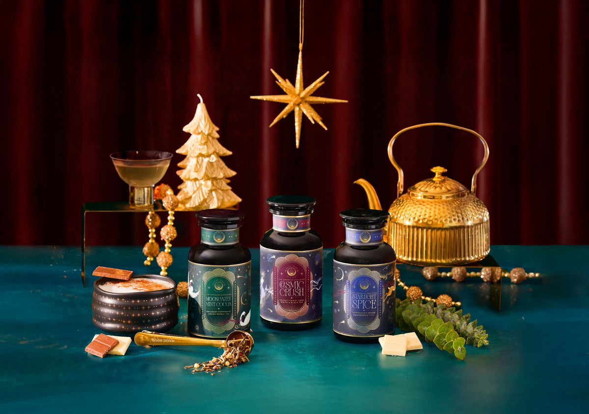 Three jars of Magic Hour tea—Moongazer Mint Cocoa, Cosmic Crush, and Starlight Spice—arranged on a teal surface with a gold teapot, a mug of cocoa, and holiday decorations.