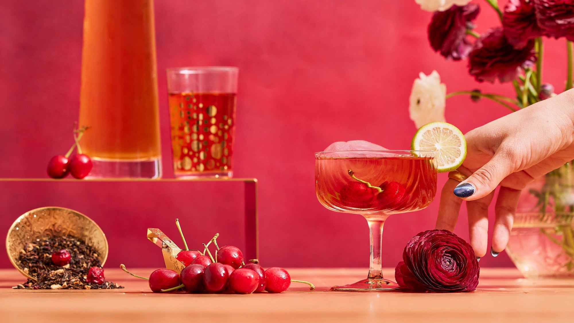 A hand reaches for a pink ranunculus flower next to a cocktail garnished with a lime slice, surrounded by cherries, loose tea, a gold spoon, and vibrant drinks against a red backdrop.