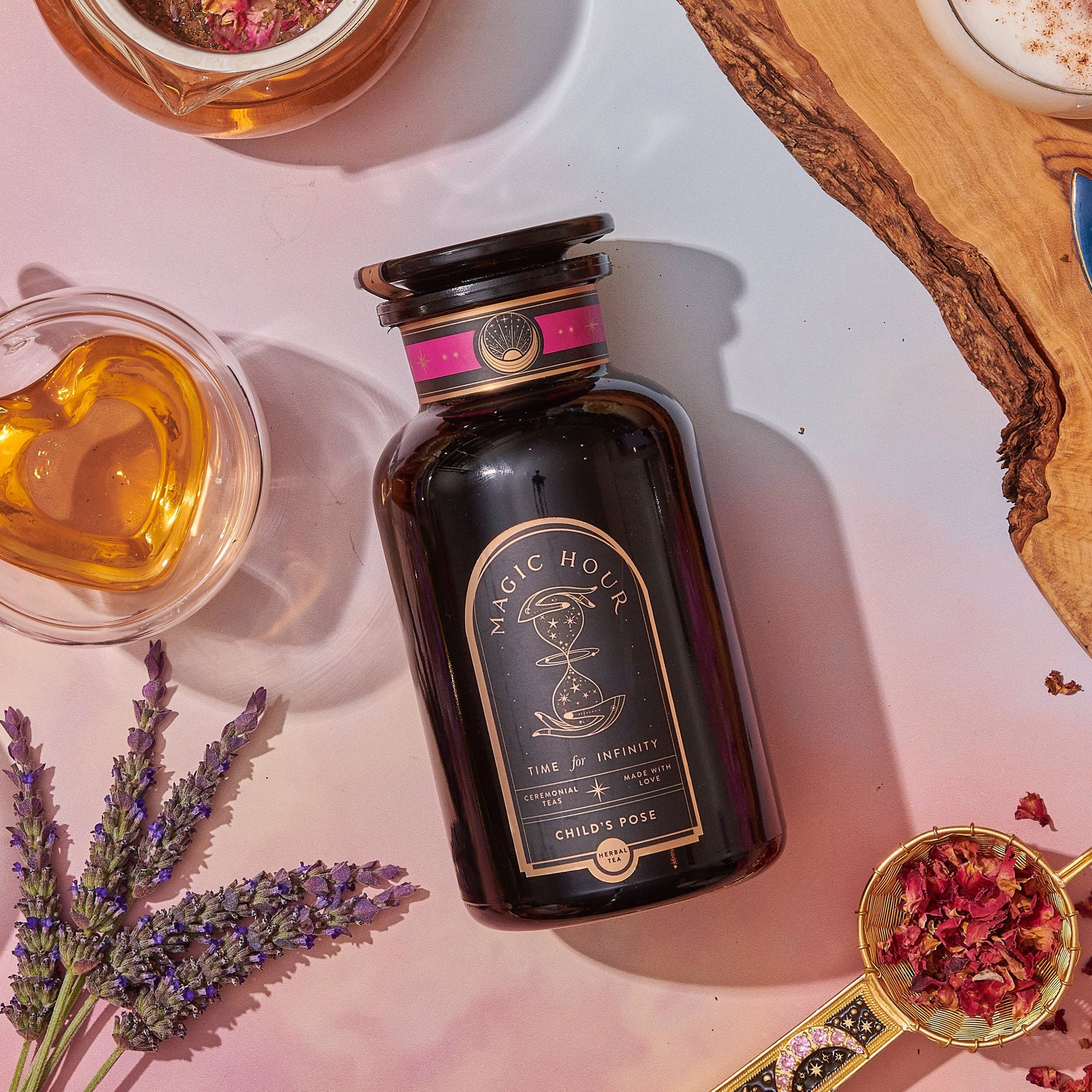 A dark glass jar labeled “Magic Hour Child’s Pose” sits on a pastel surface, surrounded by lavender sprigs, rose petals, a cup of tea, and other decorative items.