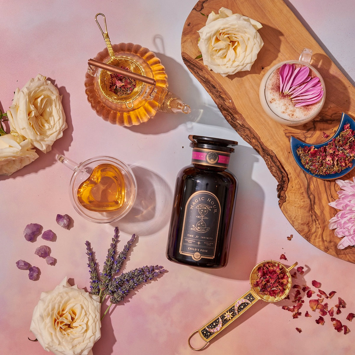 Aesthetic flat lay with a black bottle, heart-shaped glass of tea, rose petals, lavender sprigs, pink crystals, and white roses on a pink background.