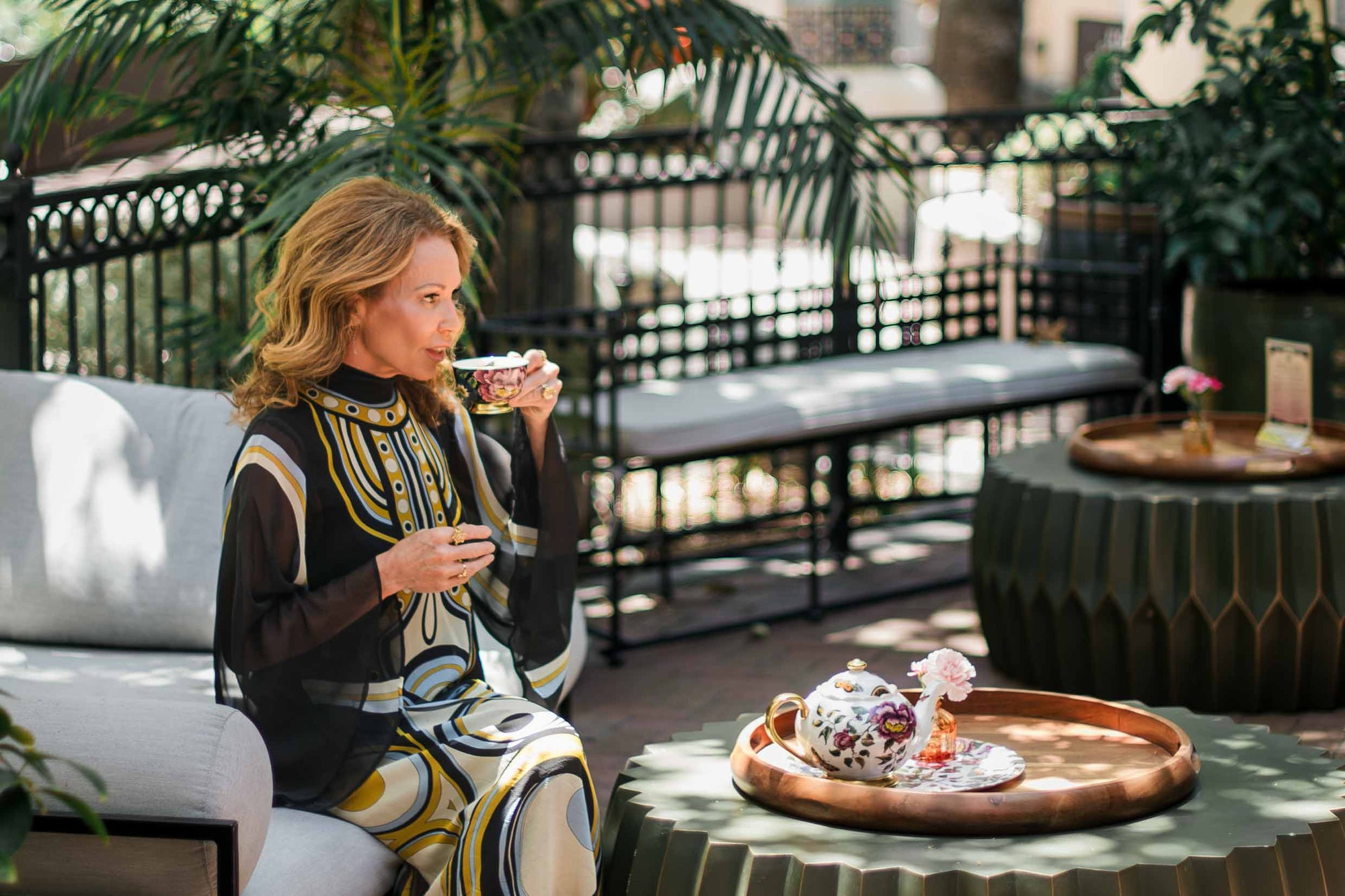 A woman in a patterned dress sits on an outdoor sofa, sipping tea from a floral cup. A matching teapot and vase with flowers are on a round table in front of her.