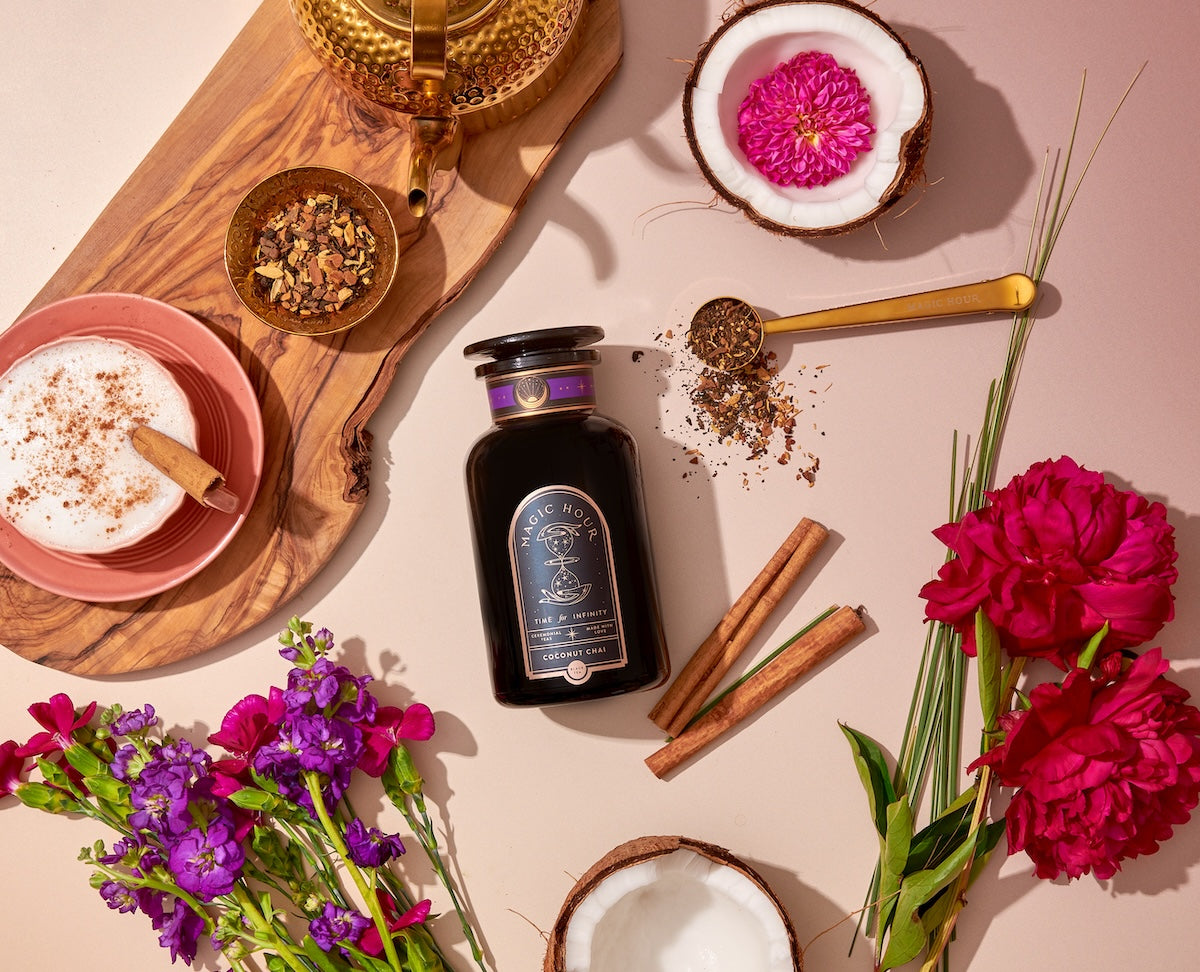 A flat-lay of a tea setup featuring a black bottle of "Magic Hour Chocolate Pu-erh," a cup of frothy tea, a golden tea strainer with tea leaves, a sliced coconut, cinnamon sticks, and vibrant purple and pink flowers.