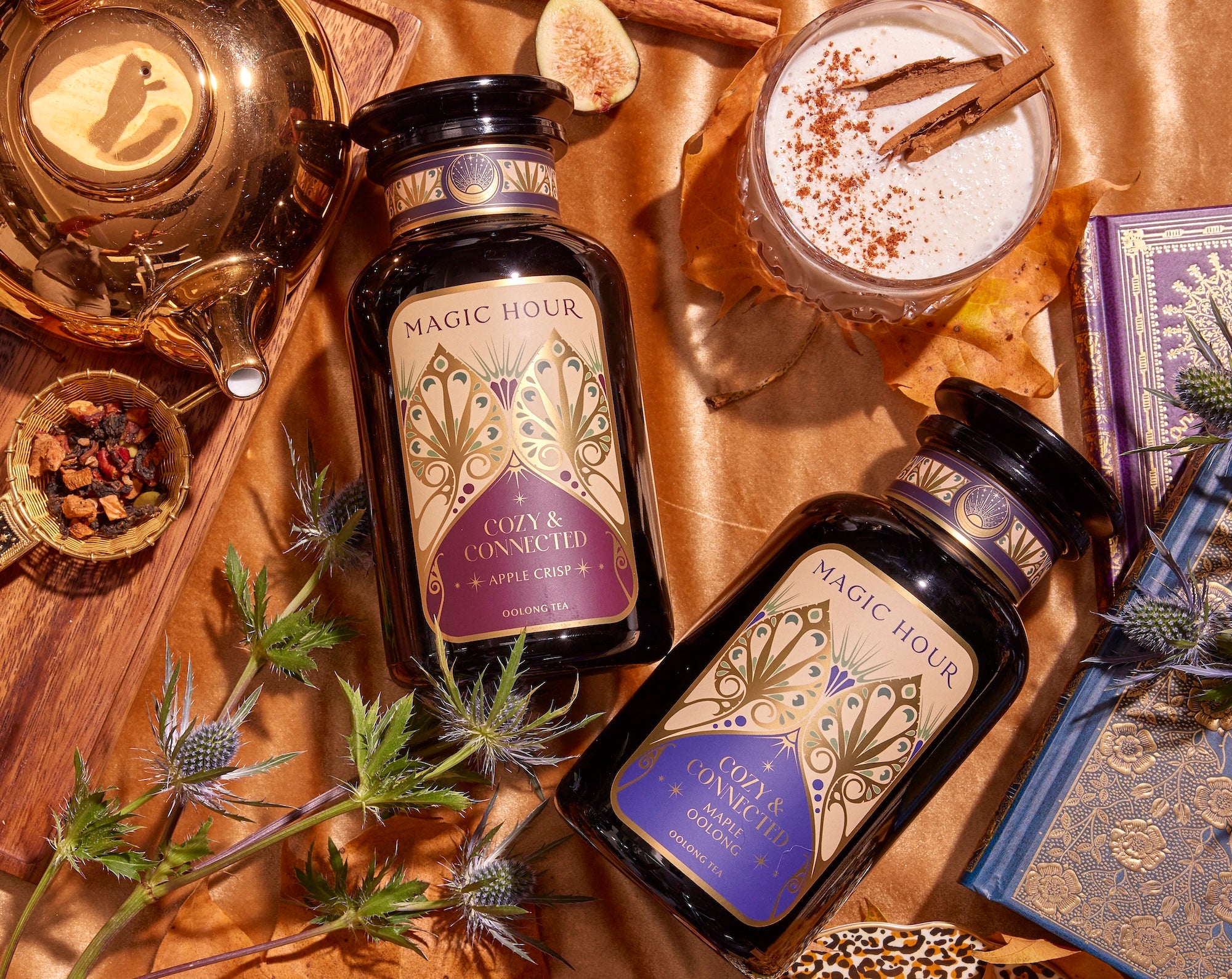 Overhead view of a cozy autumn setup featuring two jars of Magic Hour tea, a golden teapot, a spiced beverage, and several books. The scene is adorned with autumn leaves, spiky thistles, and dried fruit.