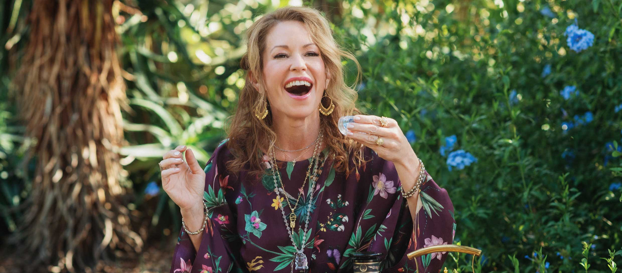 A woman in a floral dress sits outside, smiling and holding a teacup, surrounded by tea sets and greenery with blue flowers.