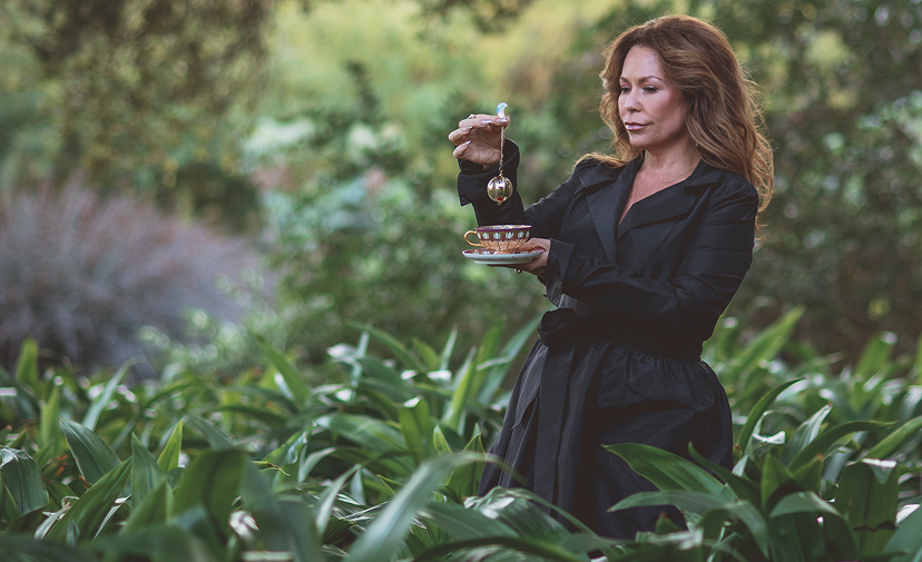 A woman in a black coat stands in lush greenery, holding a teacup and pouring tea from a teapot. The background is blurred with trees and plants.