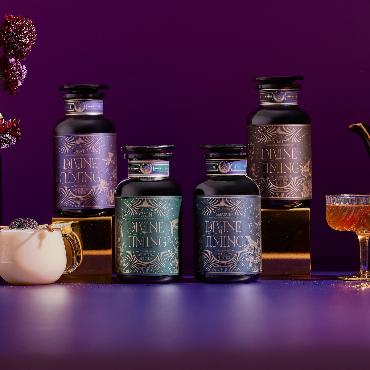A bottle of tea labeled "Queen of Cups" sits on a gold surface with tarot cards, grapes, figs, plums, berries, rose quartz, amethyst, and dried tea leaves artistically arranged around it.