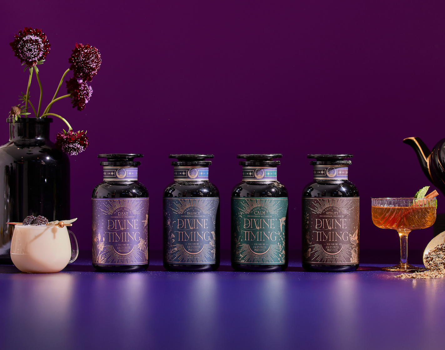 Four glass jars labeled "Divine Timing" are lined up on a purple surface with a deep purple background. Nearby are a black vase with flowers, a white mug, a teapot, and a cocktail glass with a garnish.