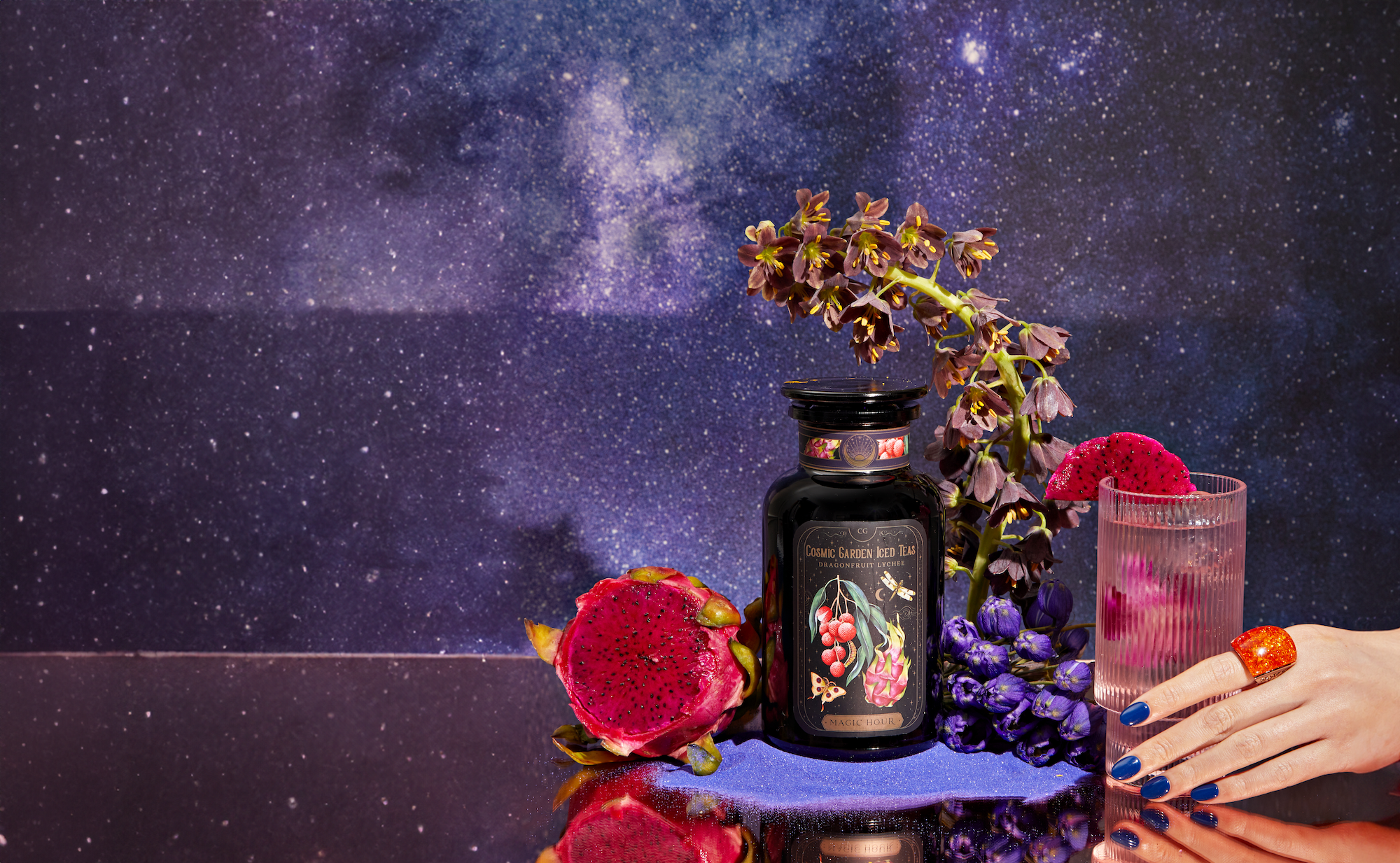 A dark jar of exotic flower and berry tea sits beside a halved dragon fruit, purple flowers, and a glass of pink drink held by a hand with blue nails, against a starry galaxy background.