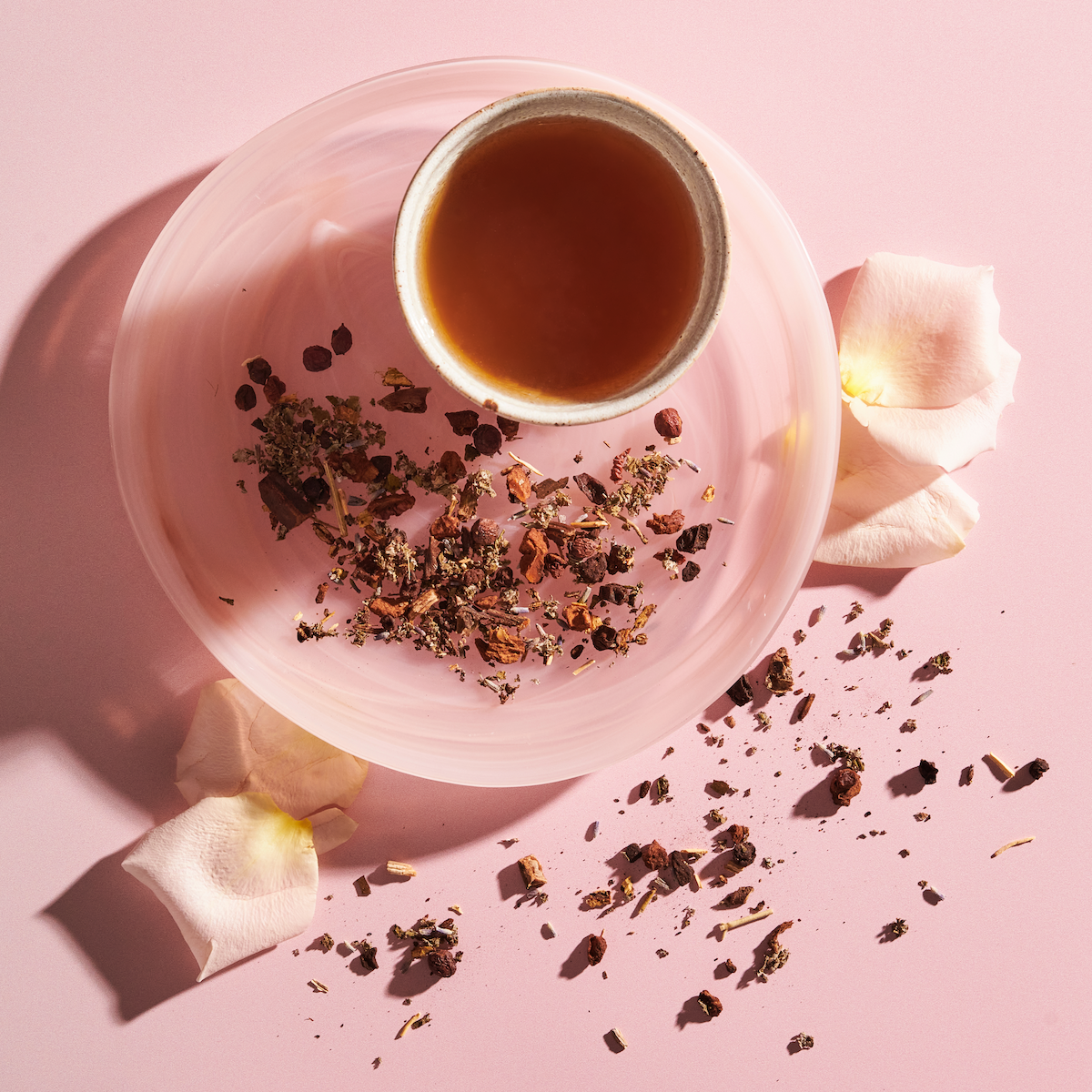 A cup of tea on a swirled pink plate with dried tea leaves and petals scattered around. The plate and cup rest on a pale pink surface. Nearby, a few pale pink rose petals add to the delicate composition.