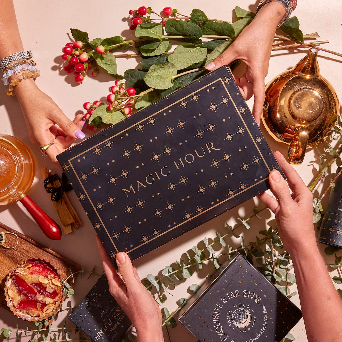 Four hands hold a black box labeled “Magic Hour,” surrounded by various tea-related items, including a teapot, honey, berries, and tea leaves on a beige surface. The scene is warm and inviting, suggesting a tea ritual.