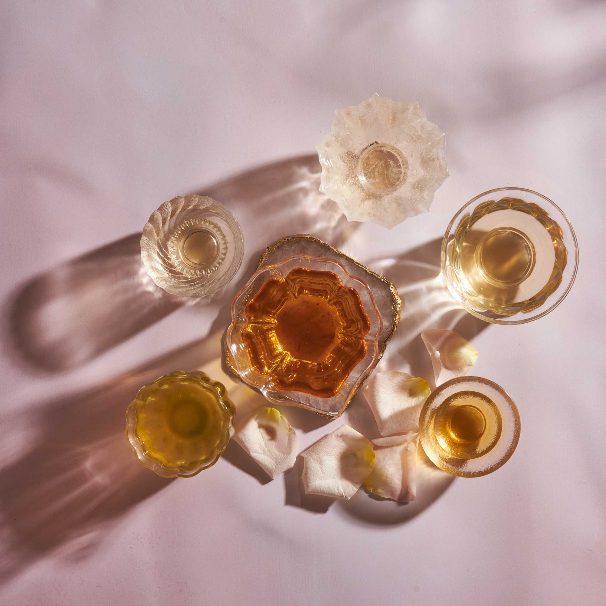 Five vintage glass cups with drinks in shades of amber and clear are arranged on a pink surface, surrounded by white flower petals and dramatic shadows.