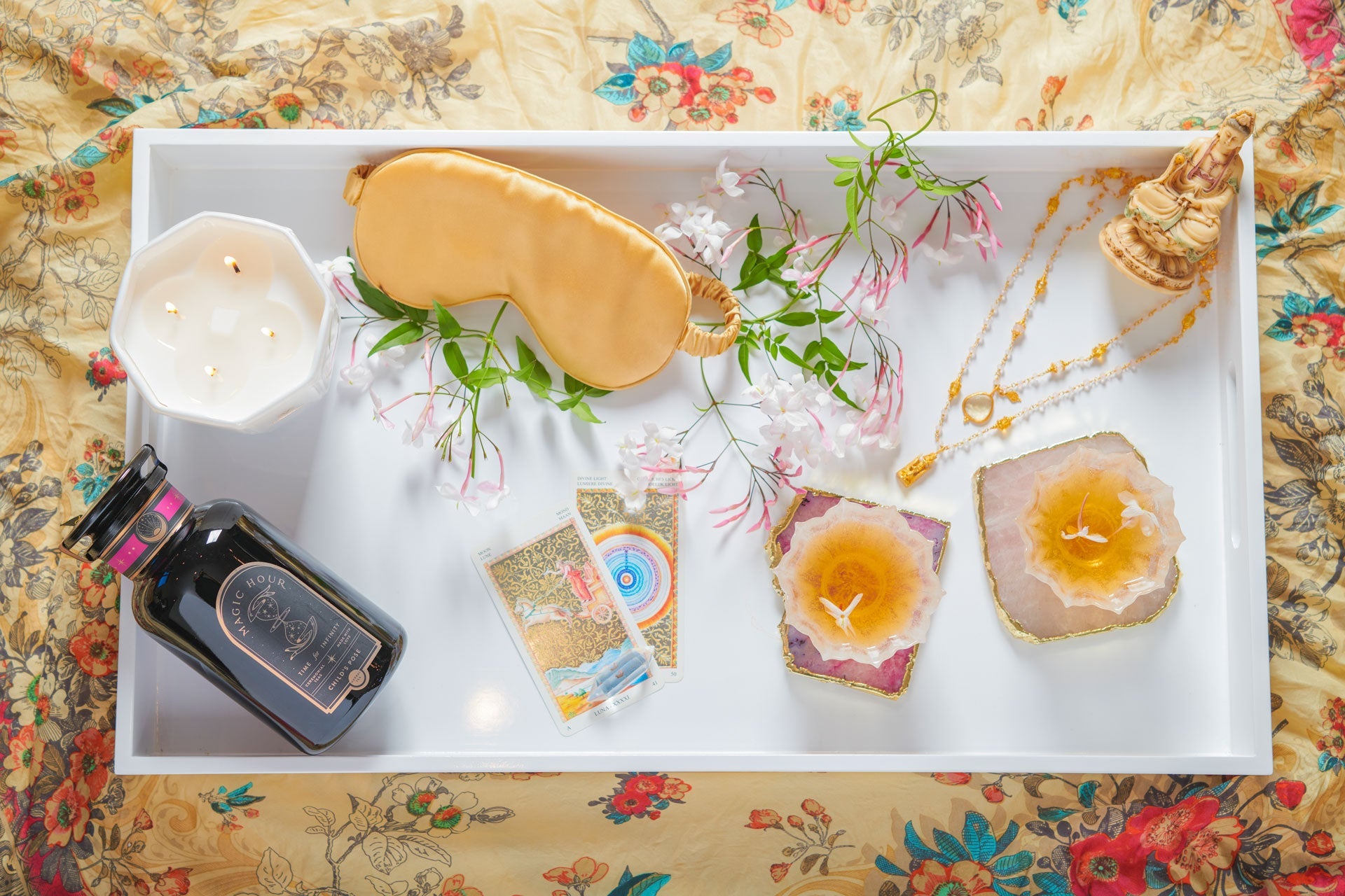 A flat lay photo of a rectangular white tray displaying two gemstone candle holders, a bottle of sleep potion, tarot cards, a sleep mask, a candle, a small golden statue, a necklace, and pink flowers, all on a floral fabric background.