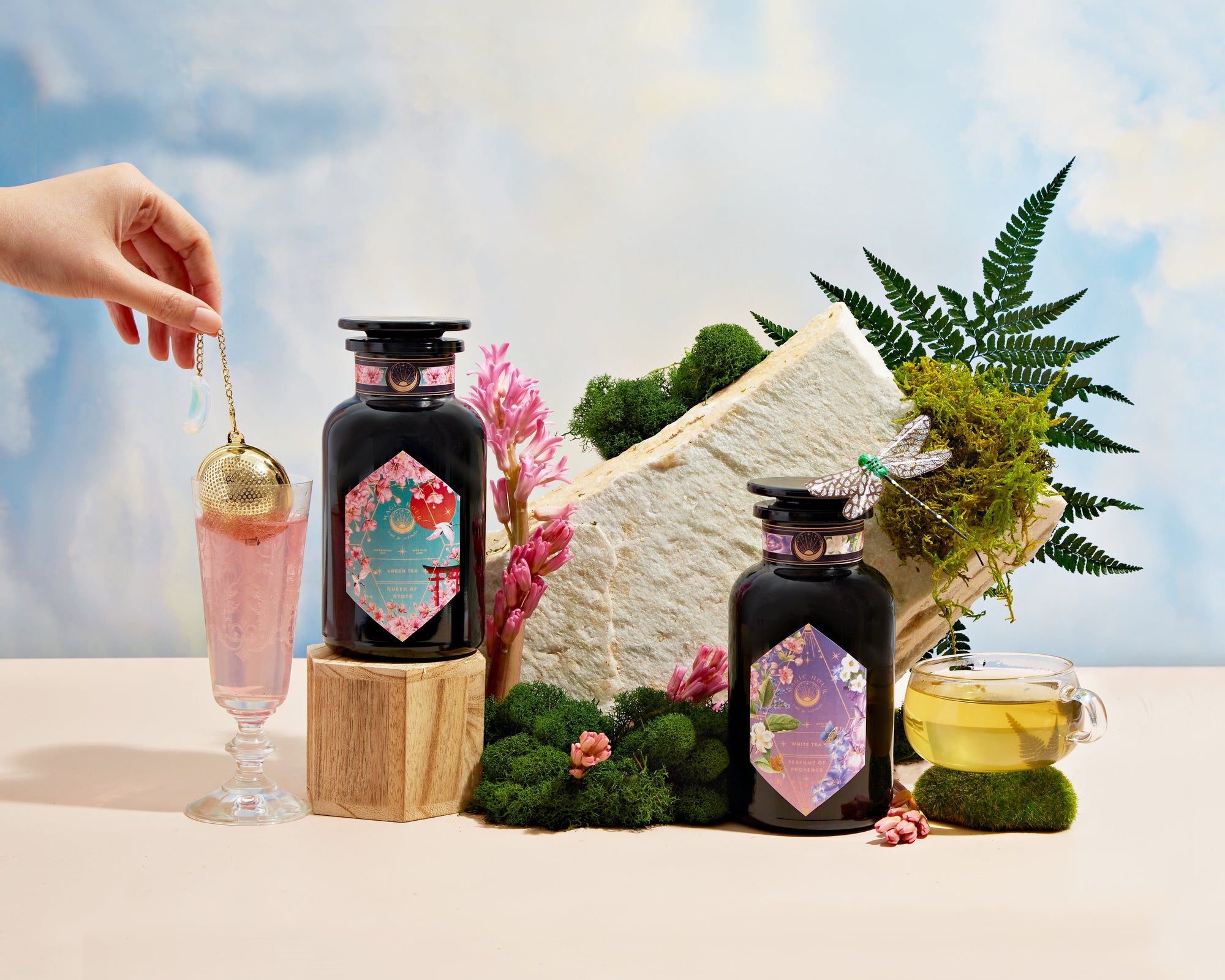A hand dips a tea infuser into a pink drink beside two decorative black jars, a teacup, green moss, flowers, and a fern against a sky-blue background.