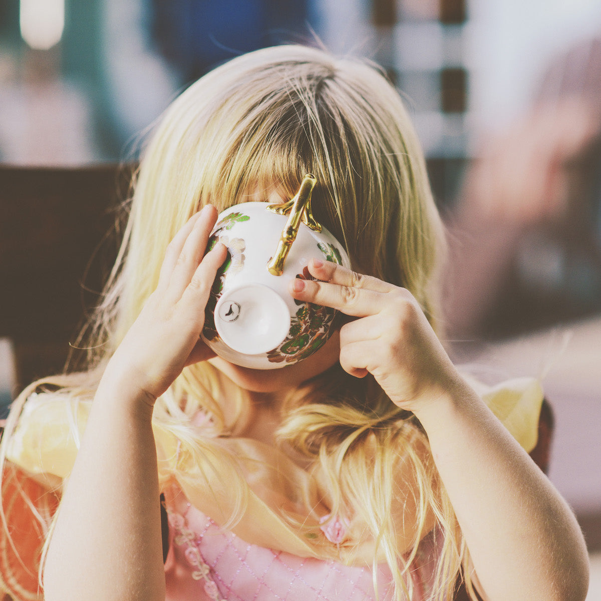 A young girl with blonde hair holds a floral teacup to her face, sipping while wearing a pink and yellow dress. She appears to be enjoying a playful tea time.