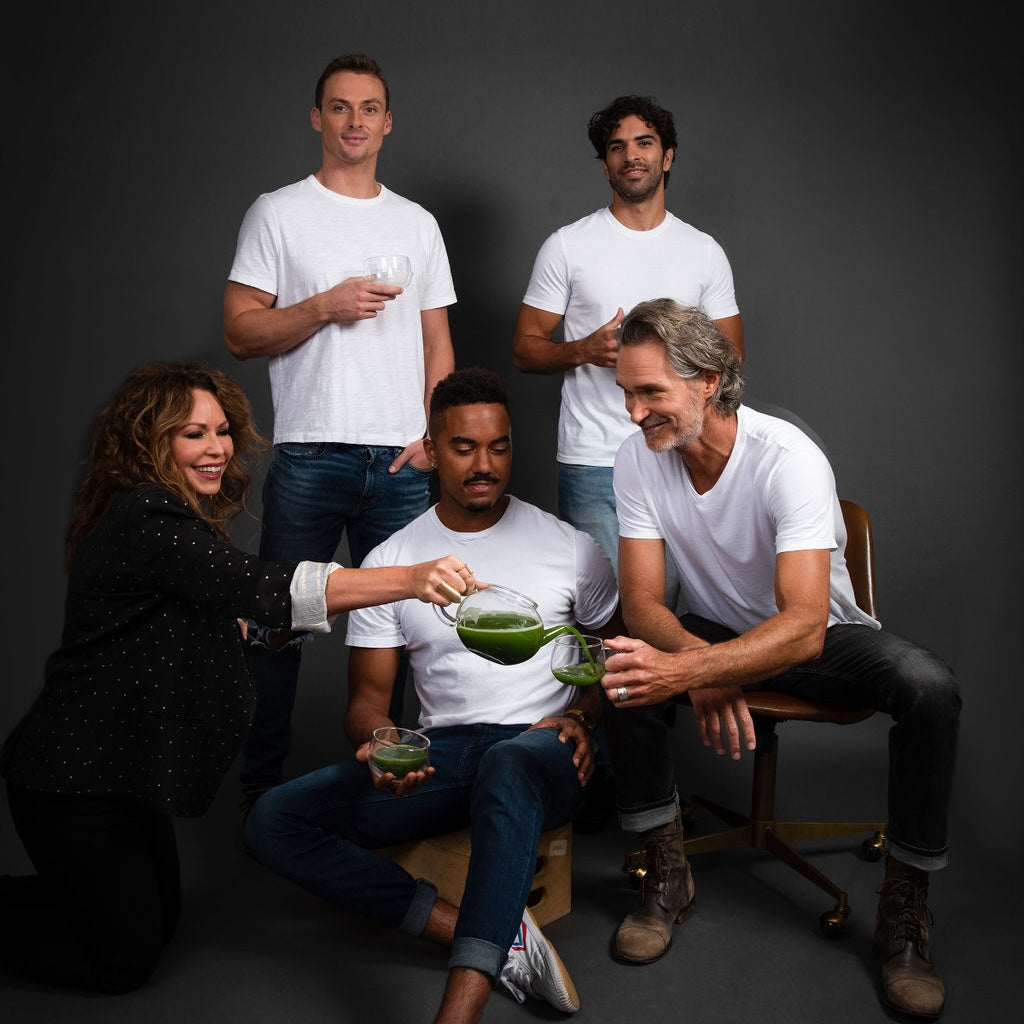 A group of five people in casual white shirts pose against a dark background. One person kneels, energetically pouring a green liquid while the others hold glasses, smiling and sitting or standing.