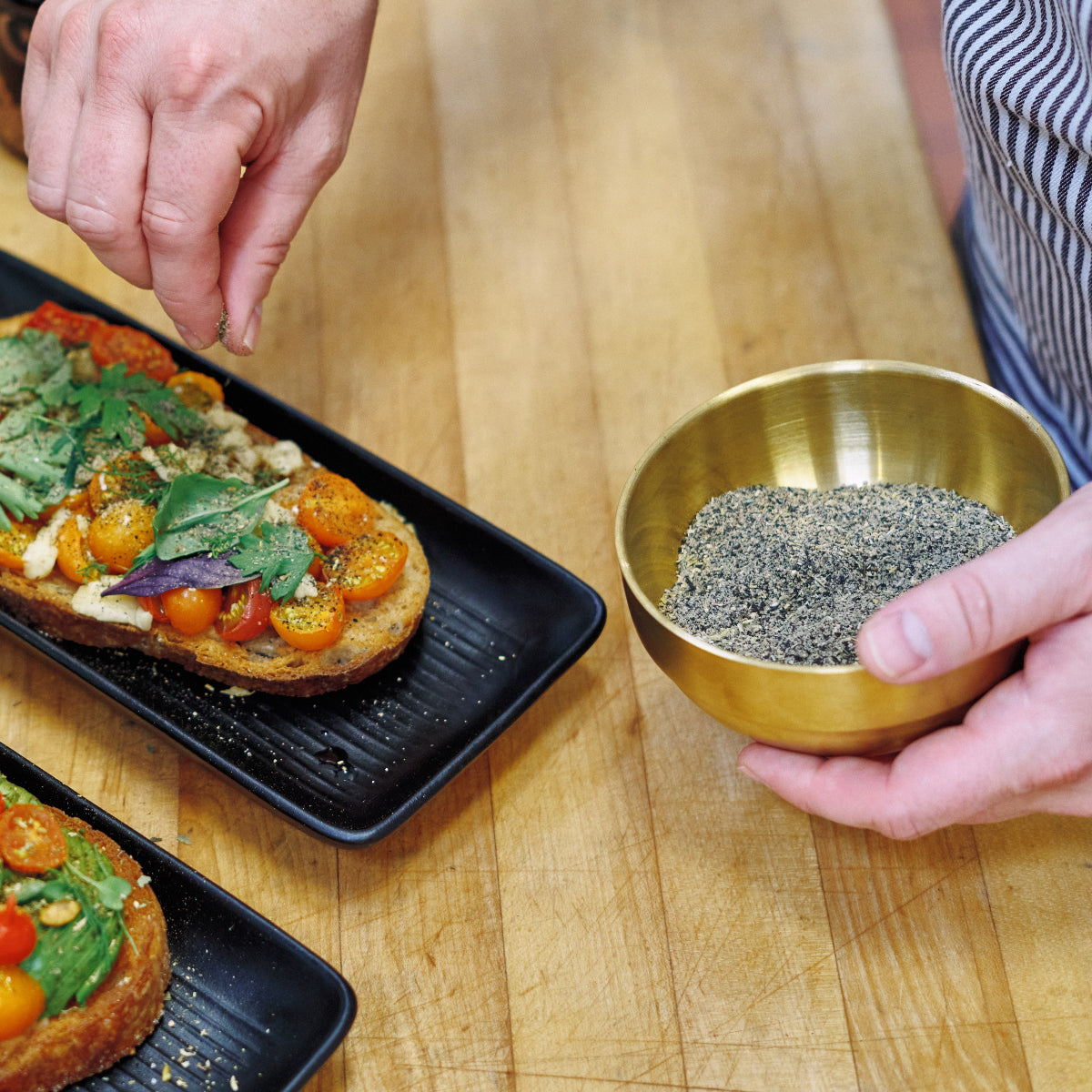 A person sprinkles seasoning from a brass bowl onto toast topped with tomatoes, herbs, and vegetables, on a wooden surface.