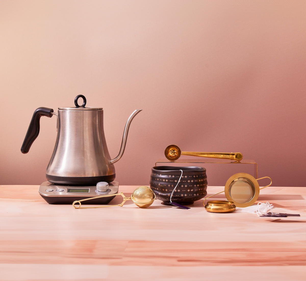 A stainless steel kettle on a digital scale, accompanied by a patterned black jar, a brass tea scoop, a round tea infuser, and a gold tea strainer, all set on a wooden table against a beige background.