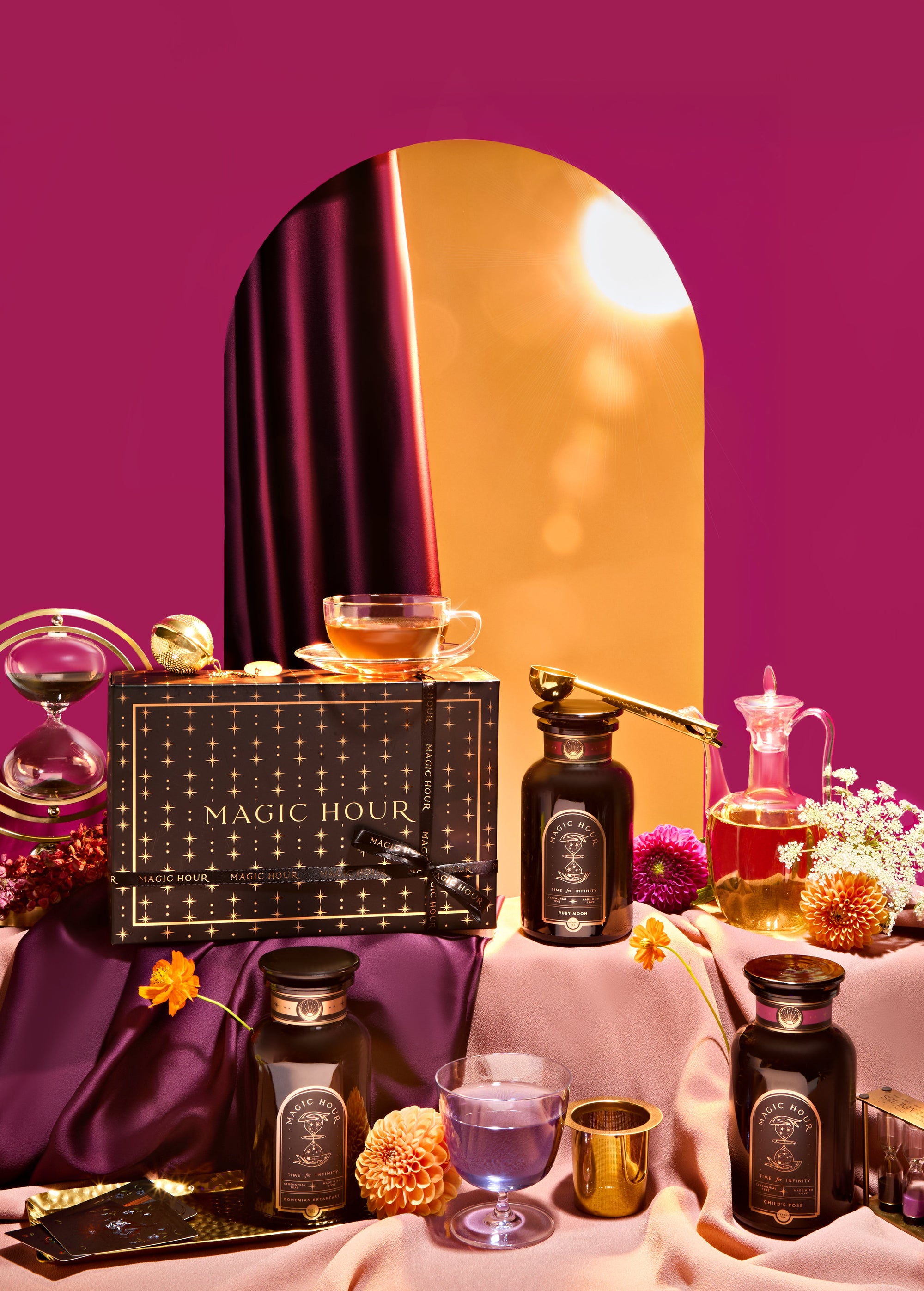 A luxurious tea set display with dark tea bottles, glasses, a cup of tea, flowers, and a "Magic Hour" gift box arranged on draped fabric, framed by a golden arch and magenta backdrop.