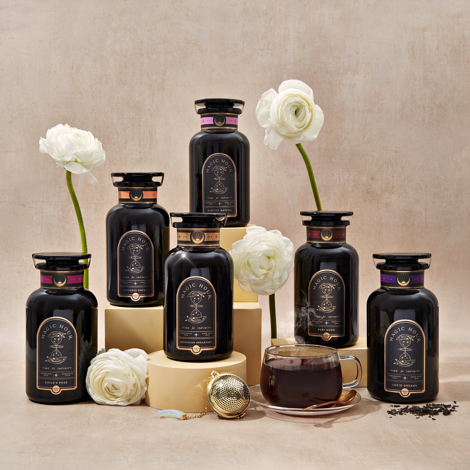 Six black tea jars with ornate labels are arranged on beige stands among white flowers. In front, a steaming cup of tea sits by a tea infuser and loose tea leaves.