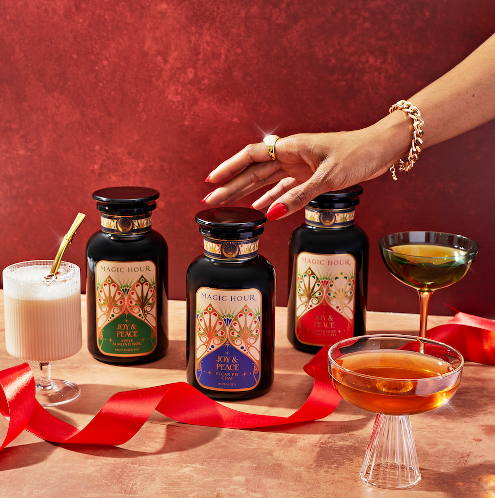 A hand reaches for one of three Magic Hour tea jars on a red surface, each adorned with floral labels. Two cocktails and a white drink in a glass with a gold straw accompany the jars, decorated with a red ribbon.