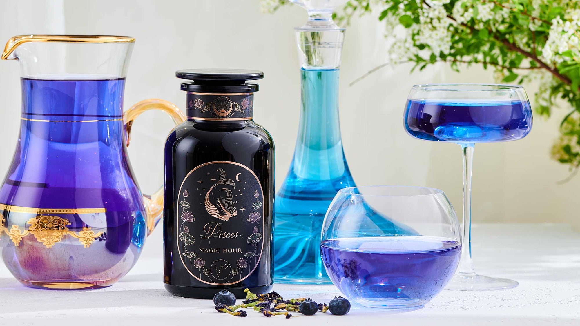A black jar labeled “Pisces Magic Hour” sits beside a glass cup of purple tea, blueberries, white flowers, and a yellow ranunculus flower against a blue background with a wavy glass panel.