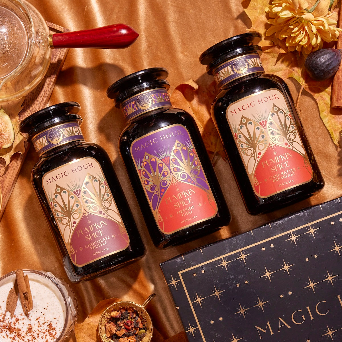 Three ornate glass jars of Magic Hour Pumpkin Spice tea are displayed on a gold satin surface, surrounded by autumn leaves, spices, a black starry box, and a creamy drink.