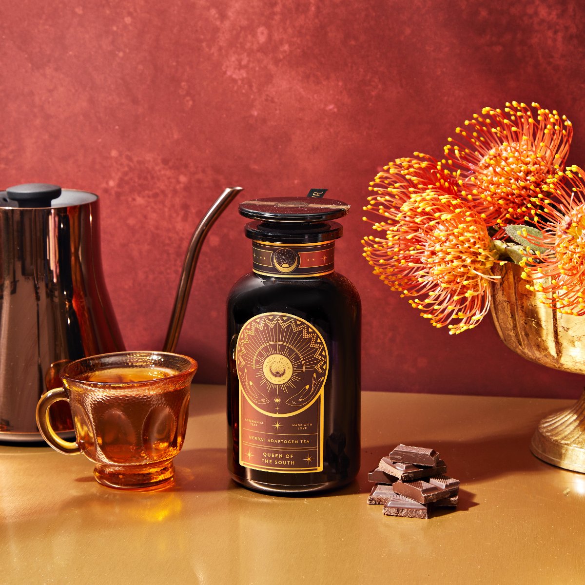 A glass teacup, a dark tea jar labeled "Queen of the South," chocolate pieces, a gold vase with orange flowers, and a metal kettle are arranged on a gold surface against a red background.