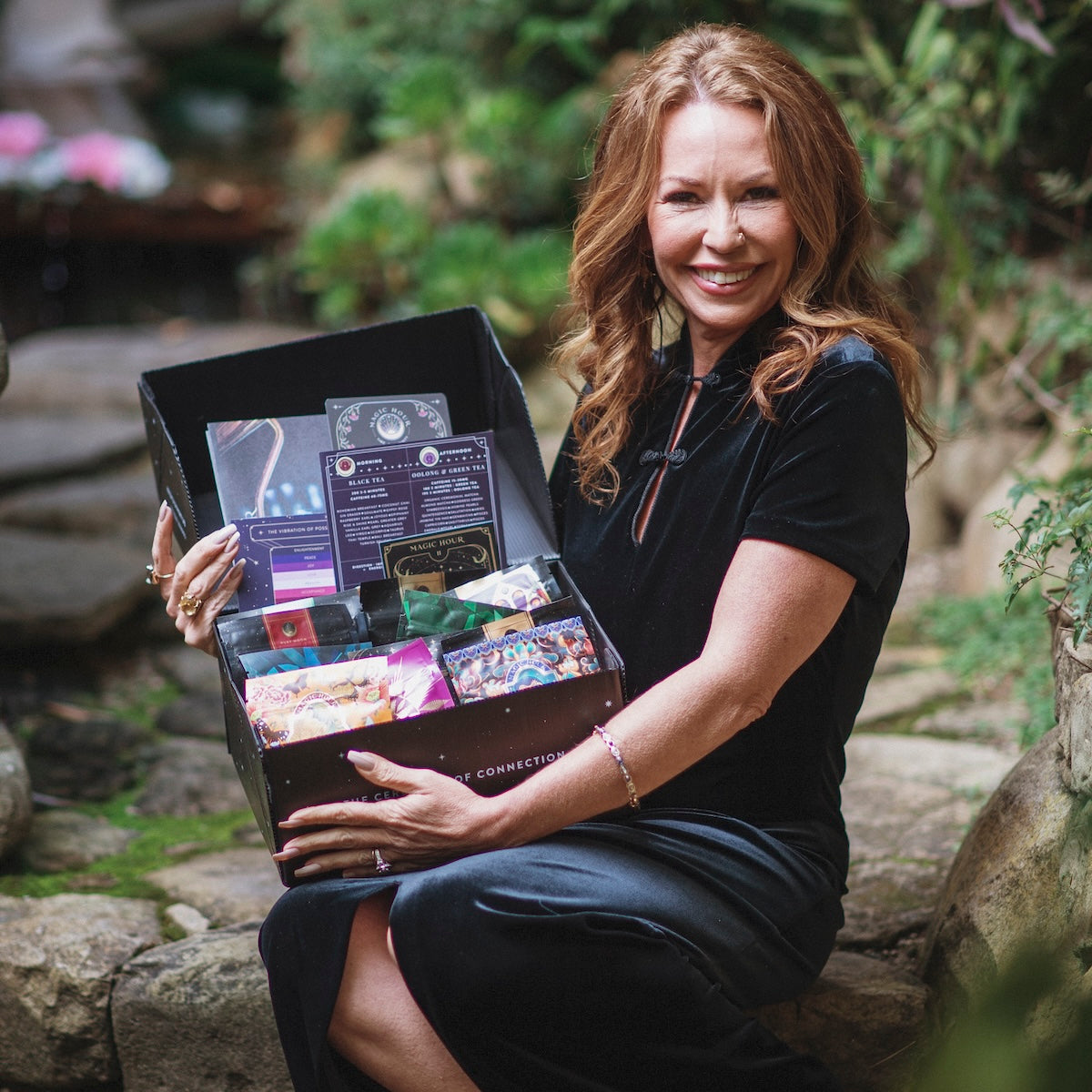 A woman with long red hair smiles while sitting outdoors, holding an open subscription box filled with various items. She is wearing a black dress and is surrounded by lush greenery.