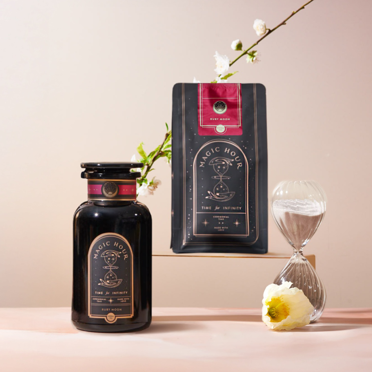 A jar and bag of Magic Hour tea with black and gold labels sit beside a glass hourglass, white flower, and blooming branch against a soft pink background.