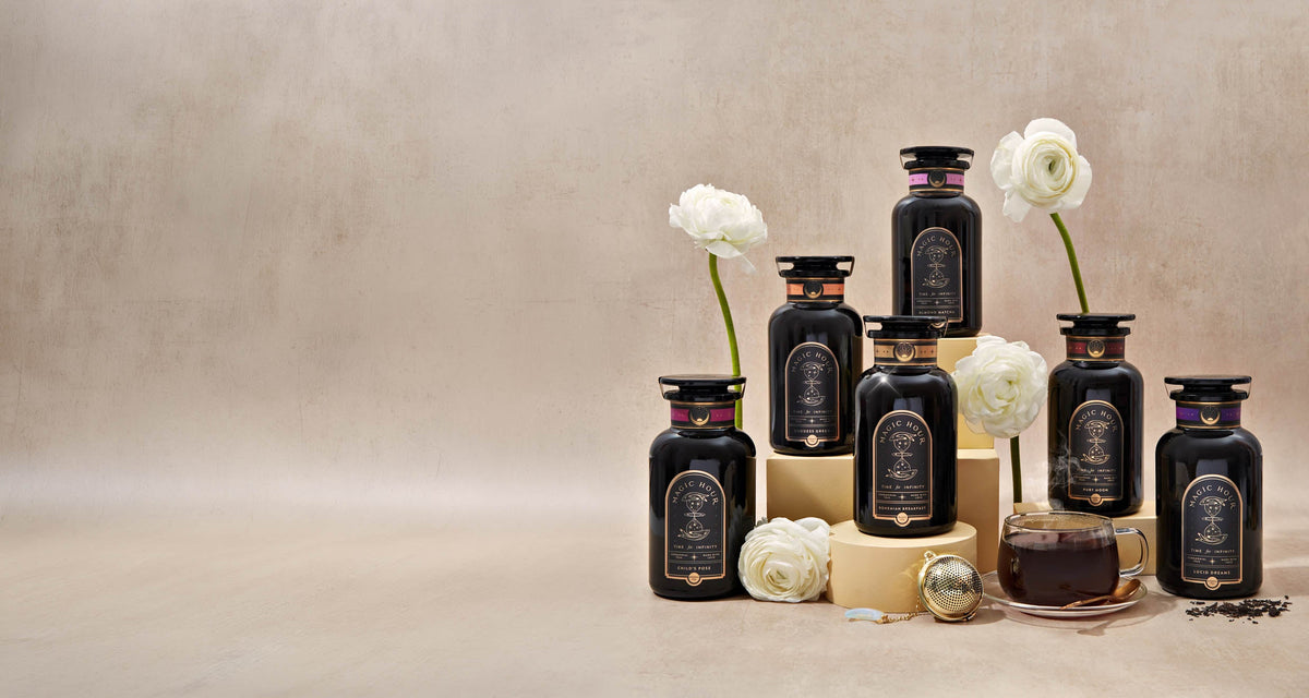 Six black jars of Magic Hour tea arranged on tiered platforms, accompanied by white flowers, a tea strainer, and a cup of tea.