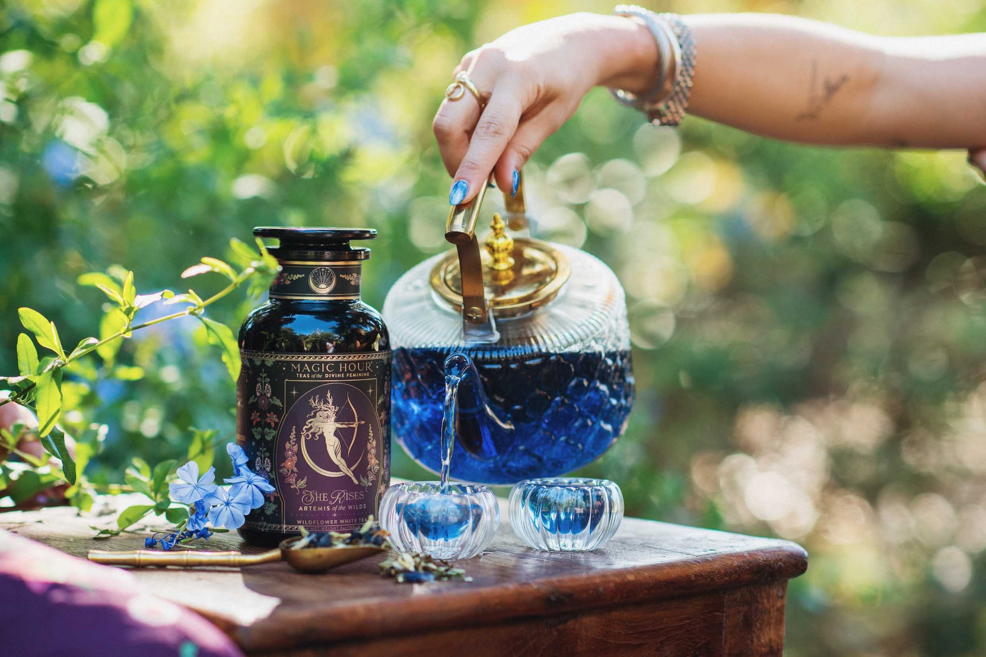 A hand pours red tea from a glass teapot into a small glass cup beside a dark floral tea canister and a whole pomegranate, all set against a lush green outdoor backdrop.