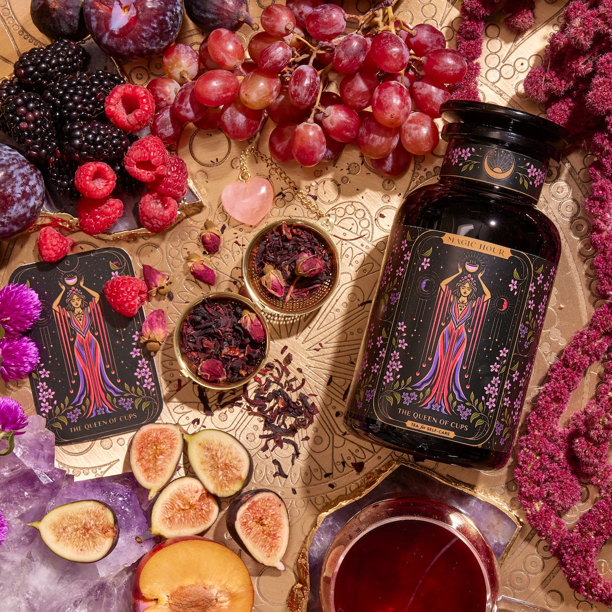 A bottle of tea labeled "Queen of Cups" sits on a gold surface with tarot cards, grapes, figs, plums, berries, rose quartz, amethyst, and dried tea leaves artistically arranged around it.