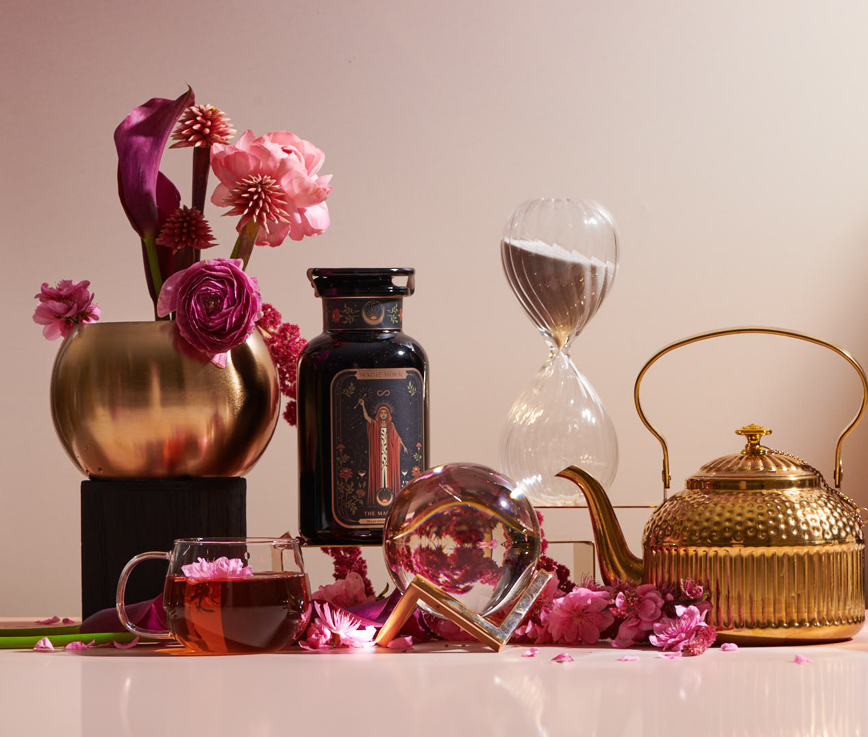 A still life with a golden teapot, an hourglass, a glass cup of tea, rose petals, and two flower arrangements among decorative black and gold items. The background is light pink, casting a warm glow over the scene.