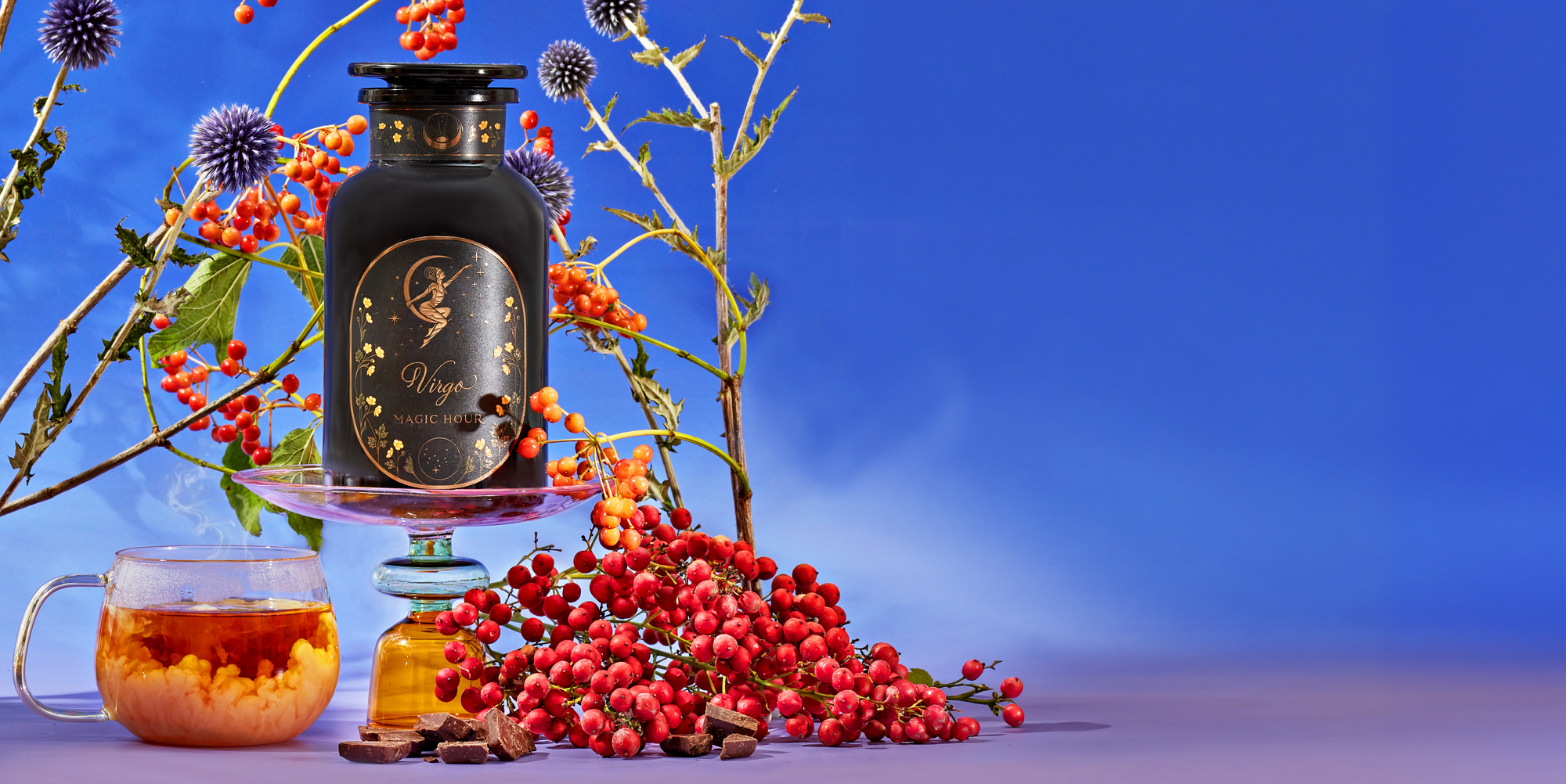 A dark jar labeled “Magic Night” sits on a cake stand among berries, herbs, a honey jar, chocolate pieces, and a cup of tea with milk, all set against a bright blue background.