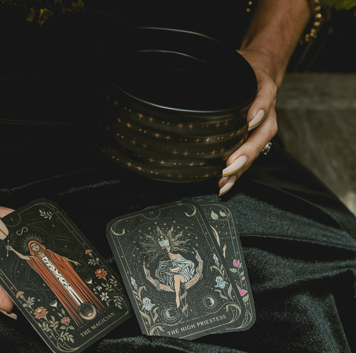 A person holds a black bowl with golden star patterns. Two tarot cards, "The Magician" and "The High Priestess," are displayed on a dark fabric background.