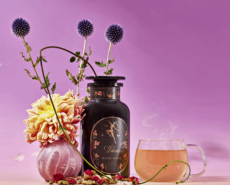 A decorative black bottle labeled "Lixir Magic Hour" is surrounded by blooming flowers and herbs on a pink background. There is also a steaming cup of tea beside it, along with scattered dried herbs or spices on the surface.